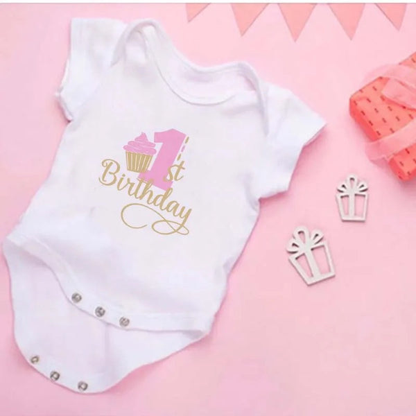 A white bodysuit with short sleeves, featuring pink cupcake graphics and the text '1st Birthday' on the front, laid flat on a pink background with decorative elements around it.