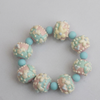 A pastel beaded bracelet featuring alternating textured round beads in pink, mint green, and cream, separated by smooth light blue beads, on a neutral light gray background.
