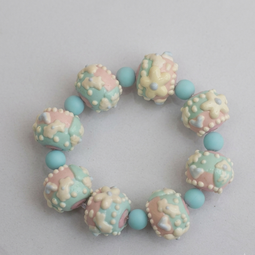 A pastel beaded bracelet featuring alternating textured round beads in pink, mint green, and cream, separated by smooth light blue beads, on a neutral light gray background.