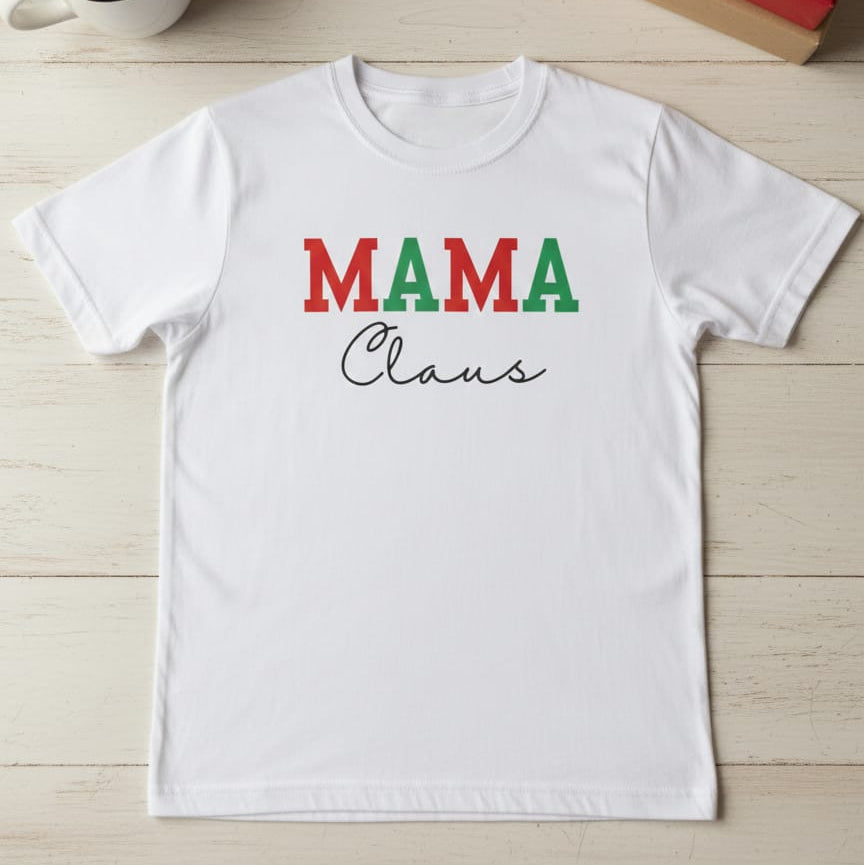 White t-shirt with 'MAMA Claus' text on a wooden surface with a cup of hot chocolate and gift box in the background.