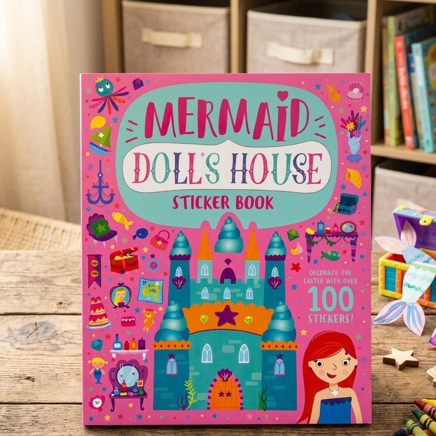 A children's sticker book titled 'Mermaid Doll's House Sticker Book' standing on a wooden table in a playroom setting. The cover is bright pink with a colorful illustration of a mermaid castle, a red-haired mermaid, and various decorative icons, plus text that says 'Decorate your castle with over 100 stickers!'.