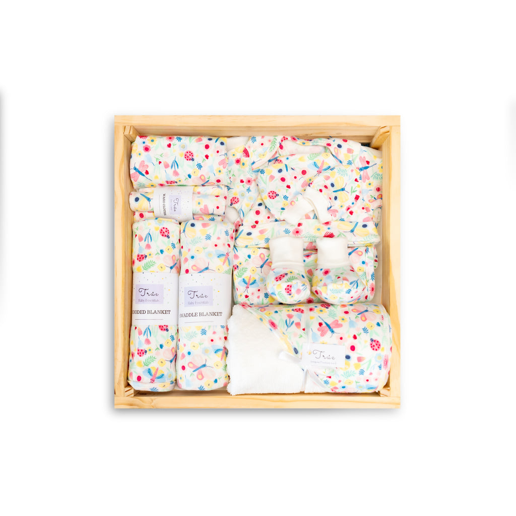 A 12-piece newborn baby essentials set in a wooden box, with a variety of items including blankets, towels, and booties decorated with a garden theme in colors like blue, pink, and white.
