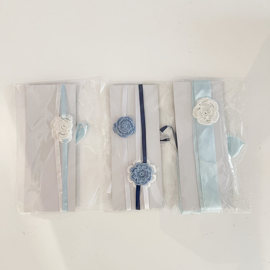 Three crochet hair bands for babies, packaged in clear plastic, featuring blue and white colors with decorative flowers and ribbons.