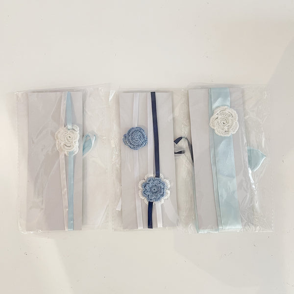 Three crochet hair bands for babies, packaged in clear plastic, featuring blue and white colors with decorative flowers and ribbons.
