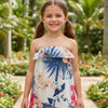 Young girl in a floral dress standing in a garden with palm trees and flowers.