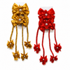 Two decorative hair bows on white backing cards, one in metallic gold and one in bright red, each with layered satin flower loops, pearl-like center accents, braided hanging cords, and small bow charms at the ends.
