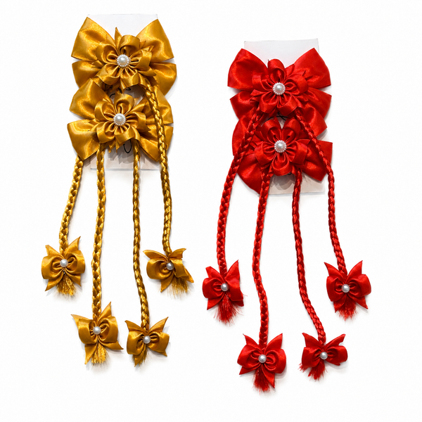 Two decorative hair bows on white backing cards, one in metallic gold and one in bright red, each with layered satin flower loops, pearl-like center accents, braided hanging cords, and small bow charms at the ends.