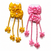 Two matching hair bows in yellow and pink, each with layered satin flower petals, pearl-like center details, braided hanging strands, and small bow accents, displayed on white backing cards against a white background.