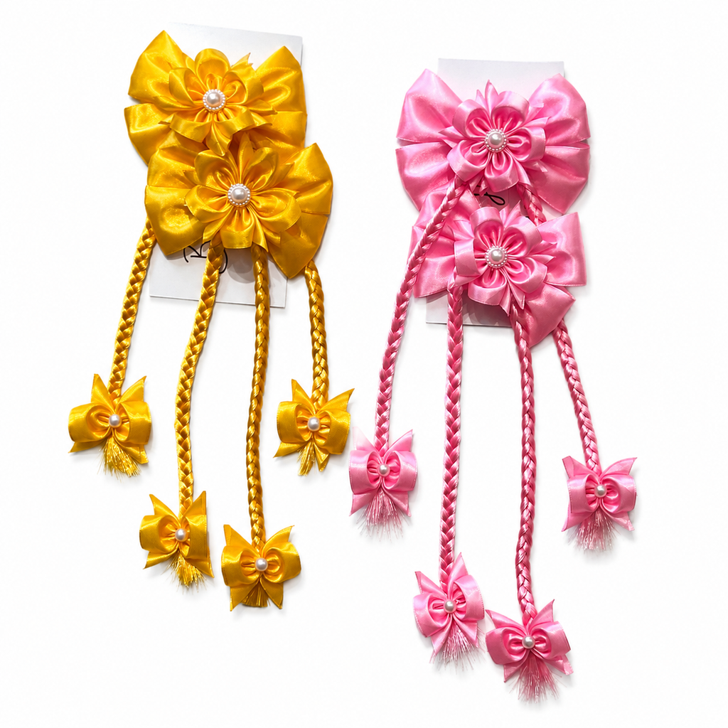Two matching hair bows in yellow and pink, each with layered satin flower petals, pearl-like center details, braided hanging strands, and small bow accents, displayed on white backing cards against a white background.