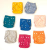 A collection of diapers in various colors, each with a padded insert visible. The colors include yellow, blue, gray, pink, and orange, all with a dotted pattern and snaps on the wings.