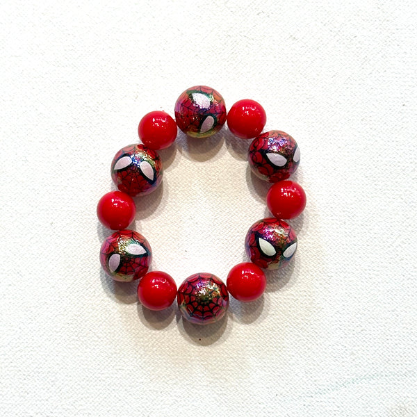 A beaded stretch bracelet featuring alternating solid red beads and spider-themed glass beads with a red and black web pattern and white spider eyes, displayed on a textured white fabric background.