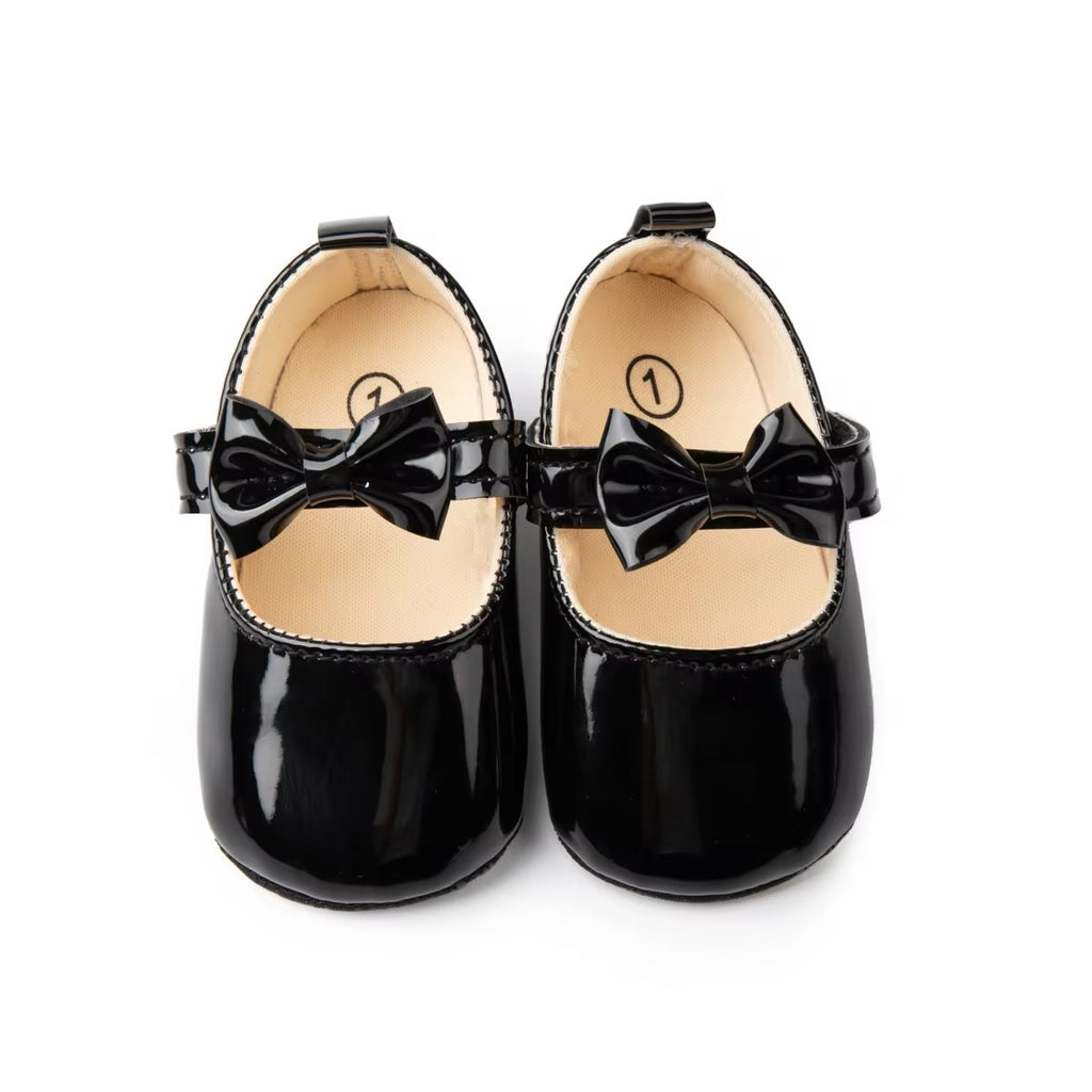 A pair of black baby shoes with a velcro strap and a bow detail on a white background.