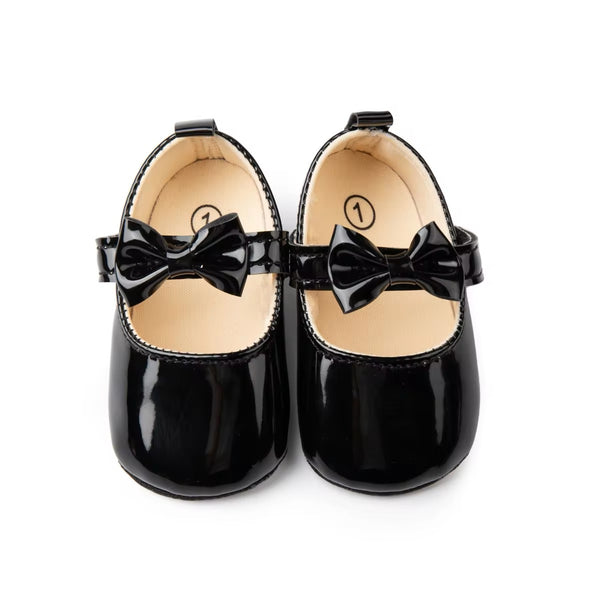A pair of black baby shoes with a velcro strap and a bow detail on a white background.
