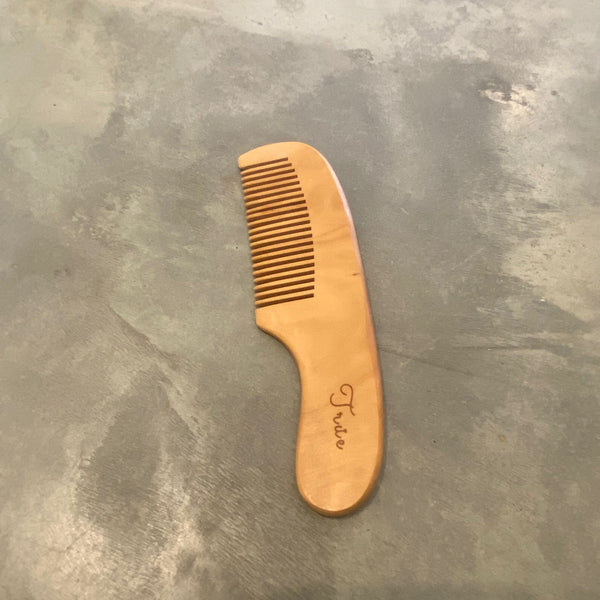 A wooden maplewood comb with rounded teeth, lying on a grey surface. The comb has a natural wood finish and the brand name 'True' is inscribed on the handle.