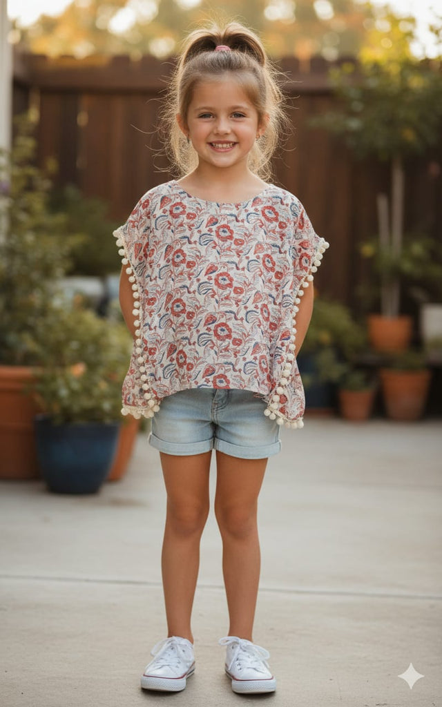 Young girl wearing a floral blouse with pom-poms and denim shorts outdoors.