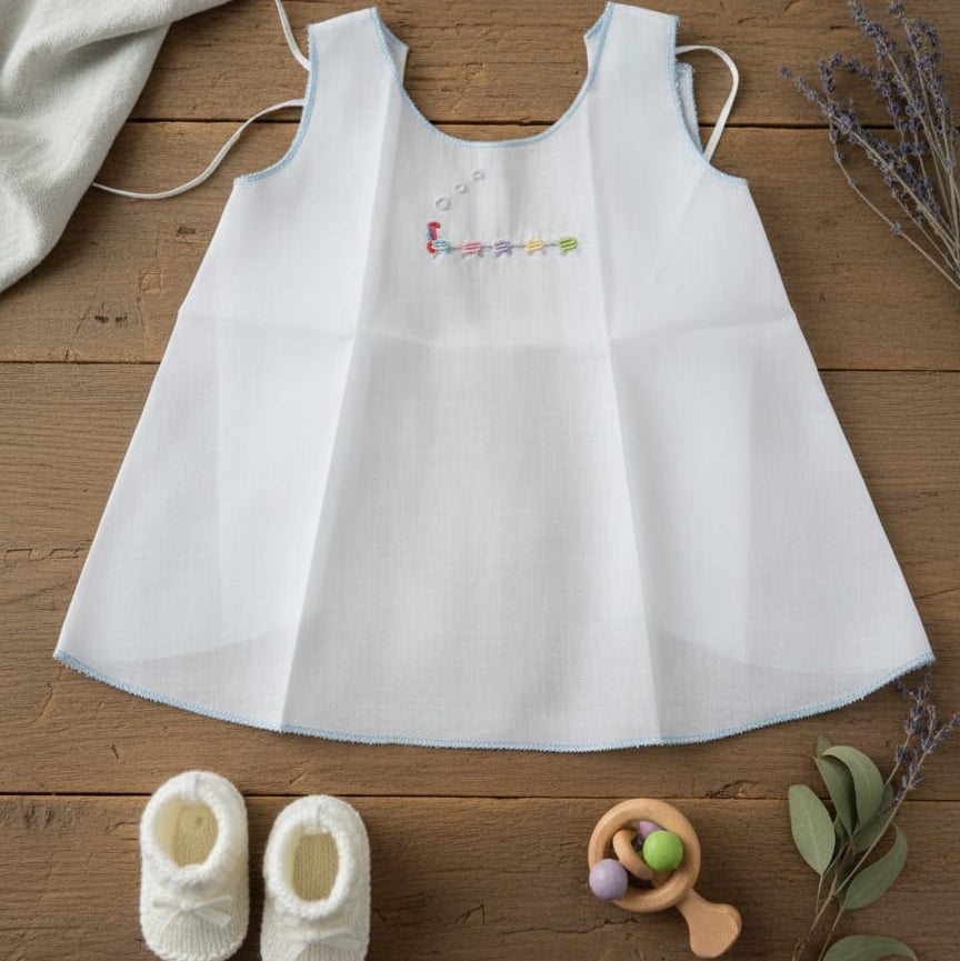 White baby dress with colorful text on a wooden surface with baby shoes and a toy.