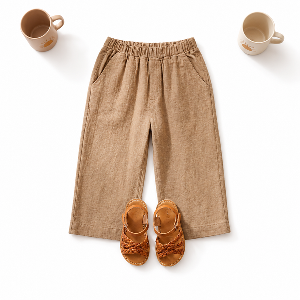 A pair of beige linen wide-leg pants with an elastic waistband, styled in a flat-lay with a pair of brown leather sandals and two ceramic mugs on a white background.