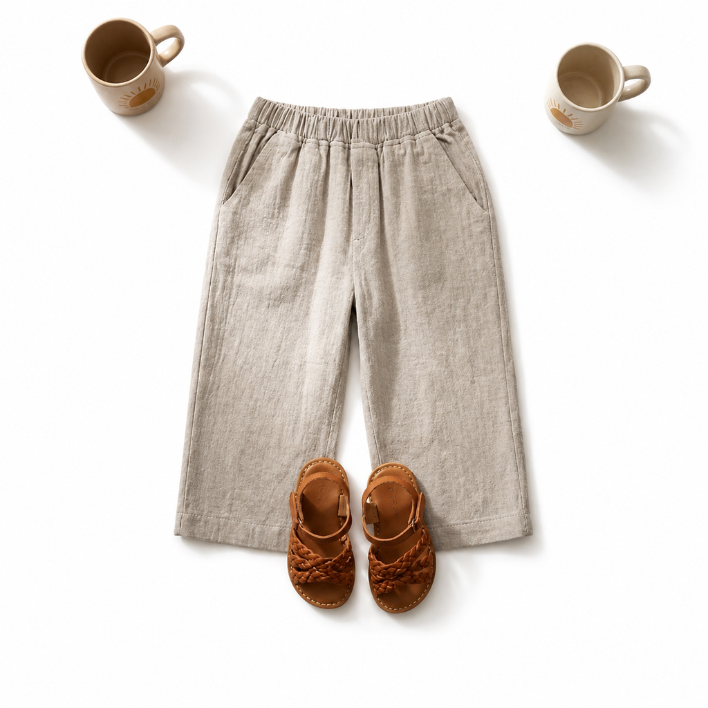 A pair of light beige linen wide-leg shorts with an elastic waistband, styled in a flat lay with a pair of brown woven sandals and two beige ceramic mugs on a white background.