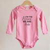 A pink long-sleeve baby bodysuit hanging on a wooden hanger against a light beige wall. The bodysuit features the text 'I'M THE Reason WE ARE late' printed in black across the chest.