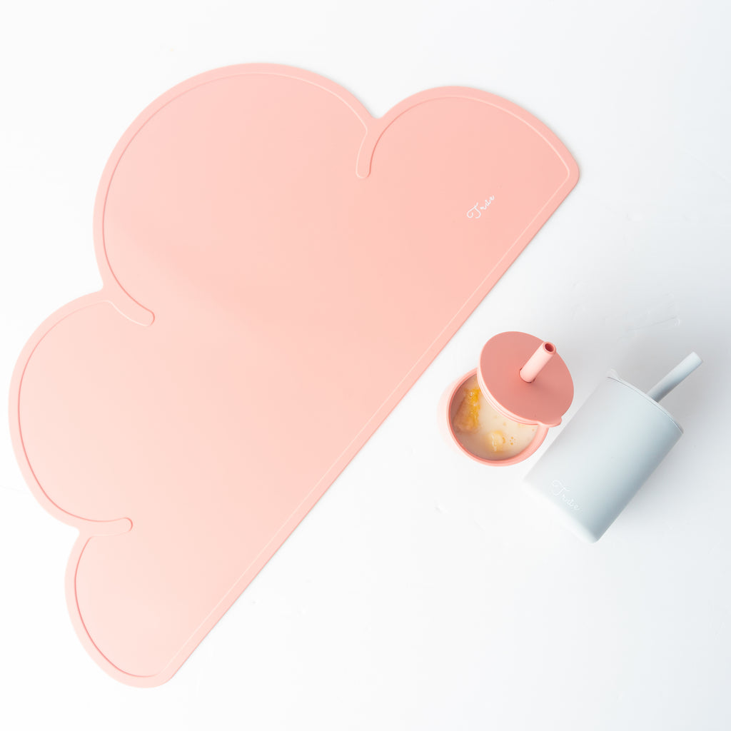 An image of a TRUE Silicone Cloud Placemat in pink, accompanied by a matching lidded cup with a pink straw and ice, as well as a blue cylindrical container, all set against a white background. The BPA-free cup complements the minimalist design of these portable items.